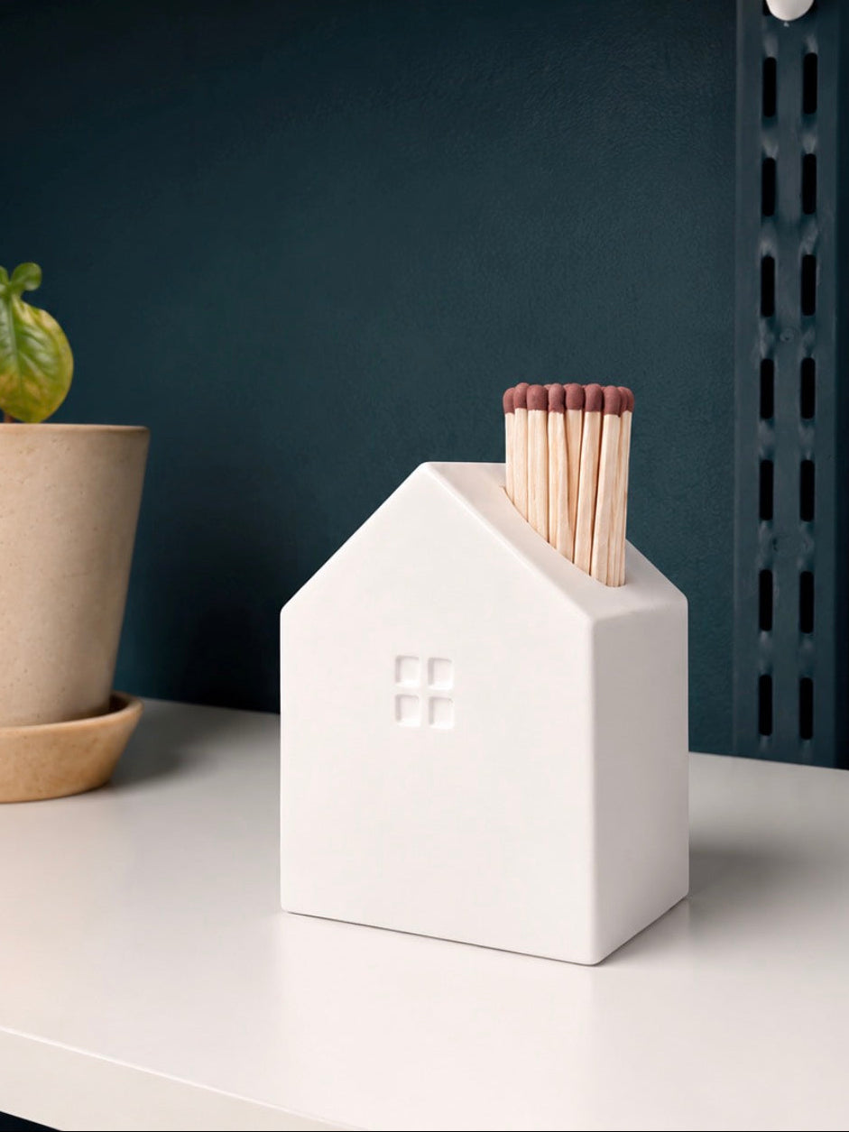 A white ceramic matchbox shaped like a small house with a window, holding wooden matches, displayed on a white shelf next to a potted plant against a dark blue wall.