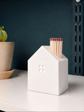 A white ceramic matchbox shaped like a small house with a window, holding wooden matches, displayed on a white shelf next to a potted plant against a dark blue wall.