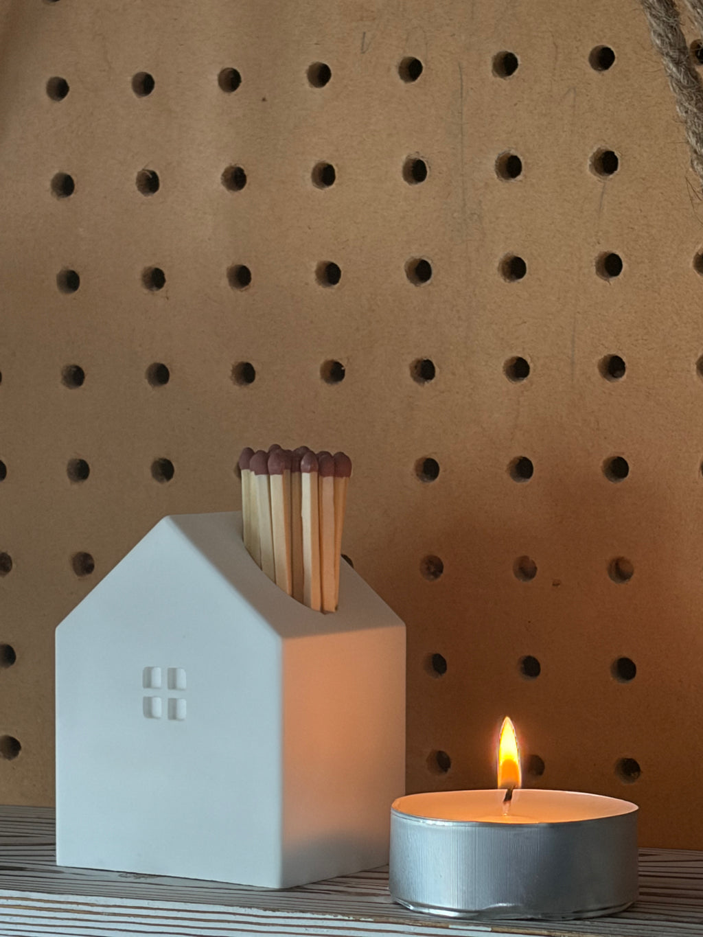 A white ceramic house-shaped match holder containing wooden matches, styled next to a lit silver tea light candle in front of a brown pegboard background.