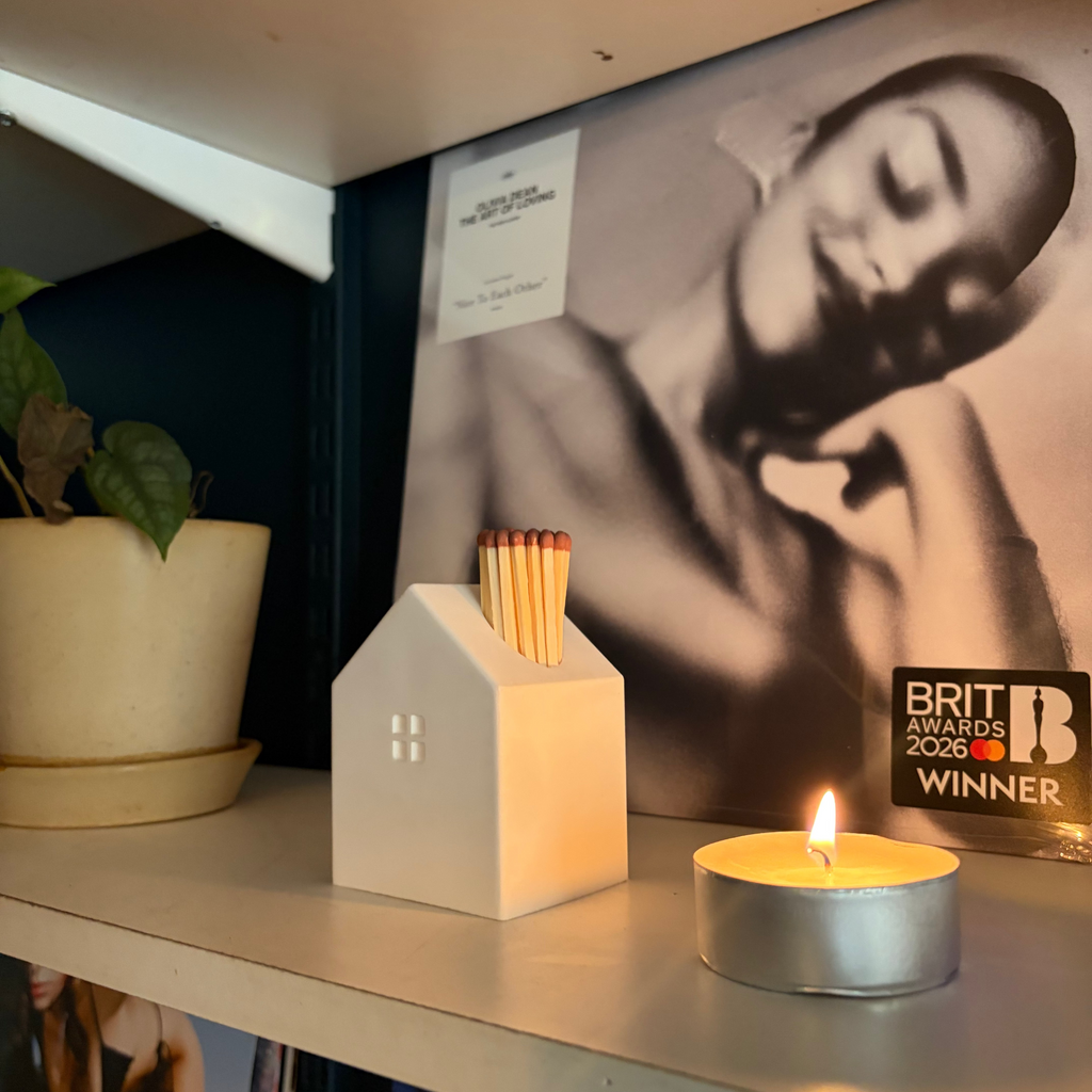 A white house-shaped match holder filled with matches sits on a shelf next to a lit tealight candle, with a black-and-white poster and a plant in the background.