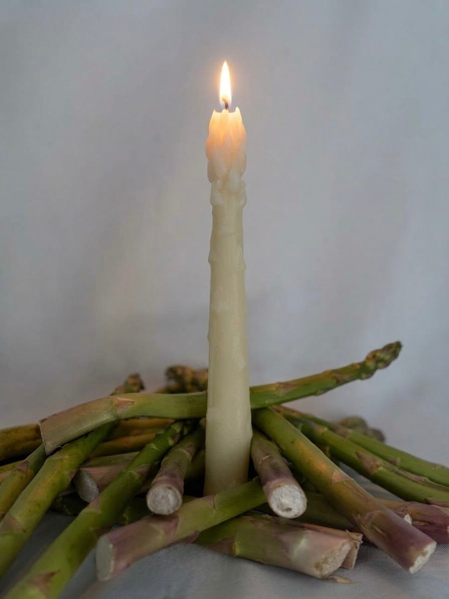Ivory beeswax asparagus taper candle lit on a bed of fresh green asparagus spears