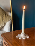 A lit white taper candle in a sculptural splat-shaped holder, set on a wooden bedside table with a dark blue headboard and a beige pillow in the background.