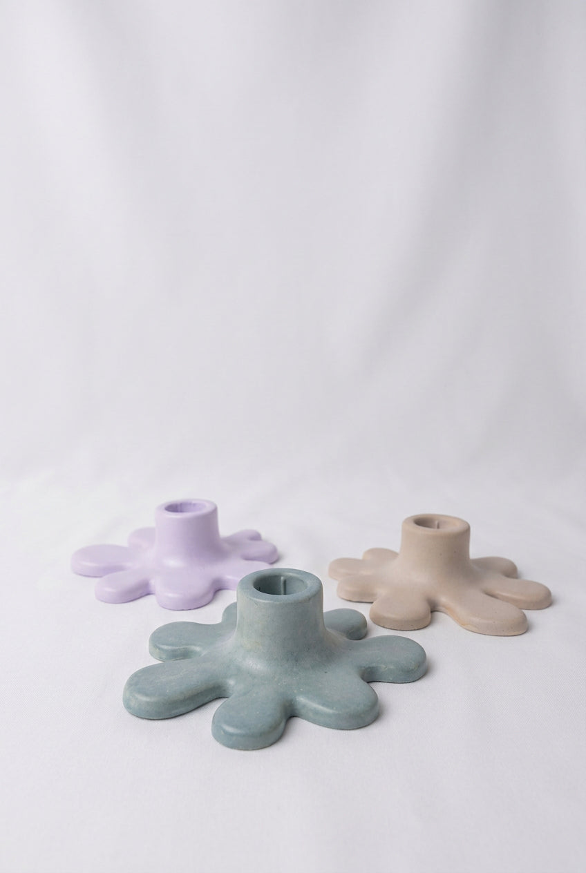 Three small splat-shaped candle holders in pastel shades of mint green, lavender, and beige , arranged on a draped white fabric background.
