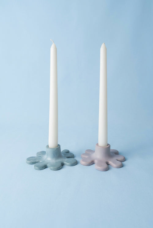 Two candlestick holders shaped like spats, one in mint green and one in light beige, each holding a white taper candle, set against a light white fabric backdrop.