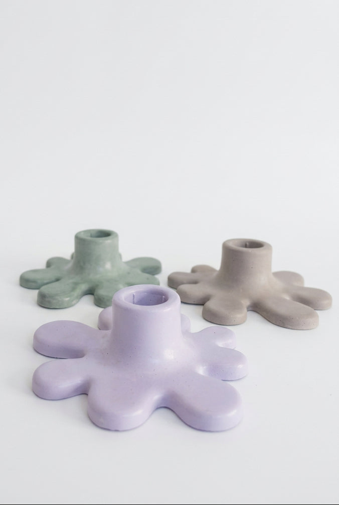 Three small splat-shaped candle holders in pastel shades of lavender, sage green, and light beige, arranged on a draped white fabric background.